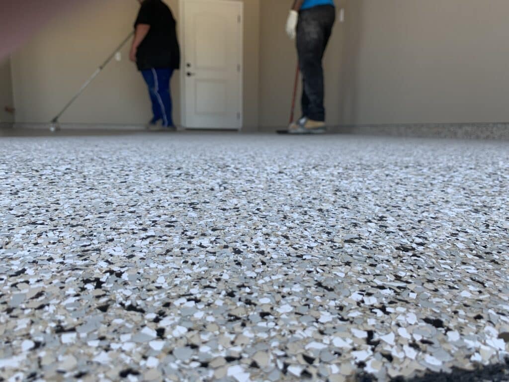 Close-up of a speckled floor with a focus on texture; two people in the background cleaning the room, one using a mop and the other stationary.