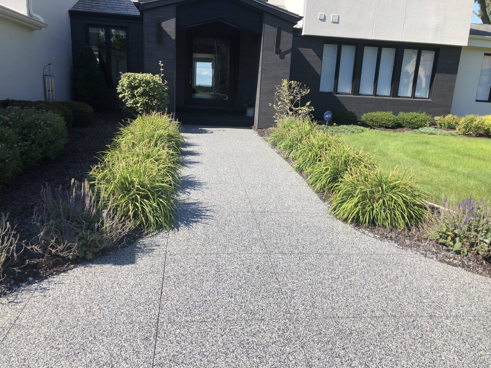 A curved concrete pathway leads to a house's front door, flanked by well-maintained garden beds with ornamental grasses and flowering plants.